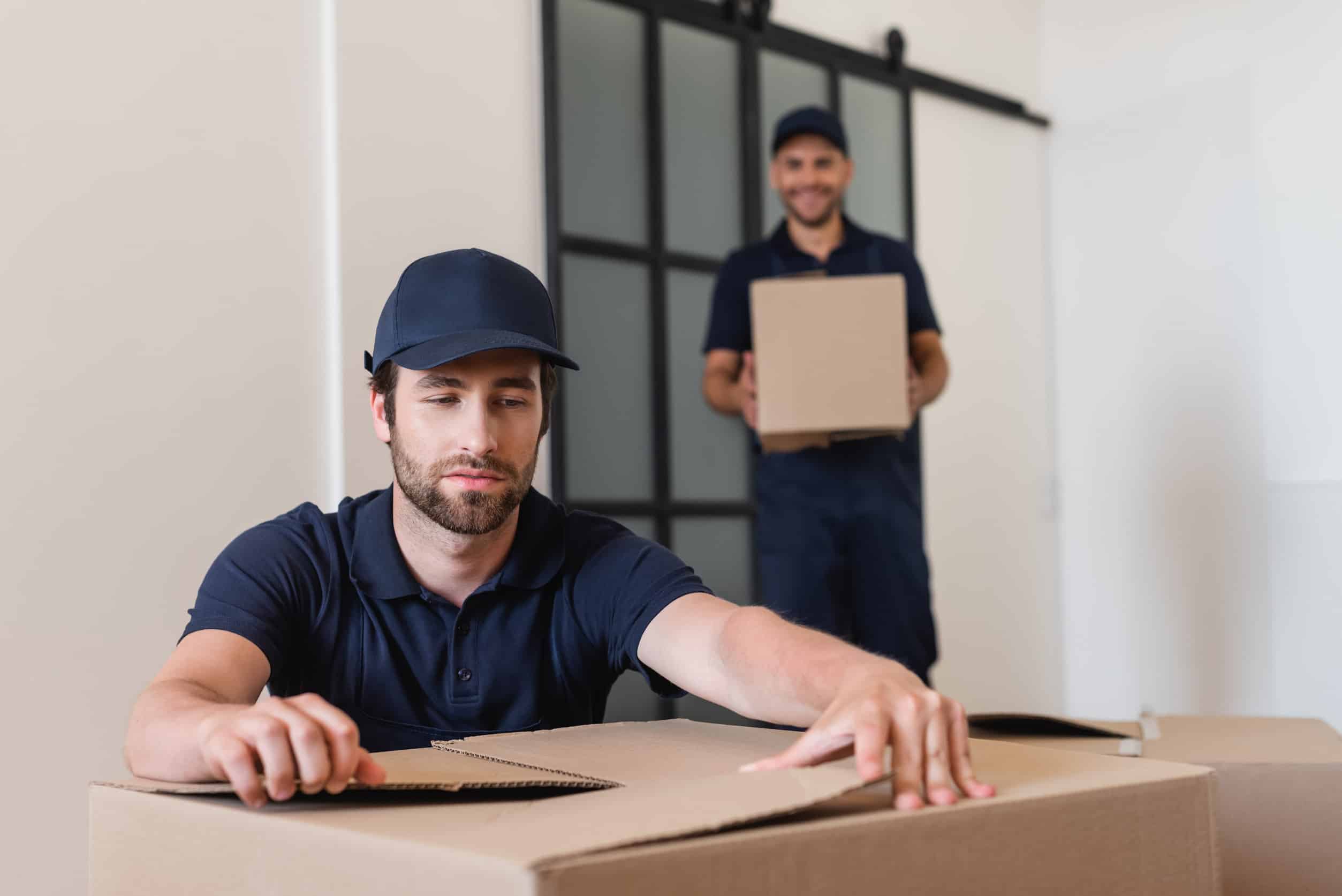 Workman in cap unpacking carton box near blurred colleague